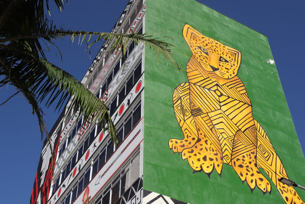 Fachada do Museu das Culturas Indígenas, em São Paulo. A imagem mostra um edifício alto com um grande mural na lateral, de fundo verde, representando uma onça estilizada em tons de amarelo e preto, com padrões geométricos. Ao lado, vê-se a estrutura do prédio com janelas e elementos arquitetônicos, enquanto uma palmeira aparece em primeiro plano. O céu azul ao fundo destaca o contraste das cores do mural e da construção.