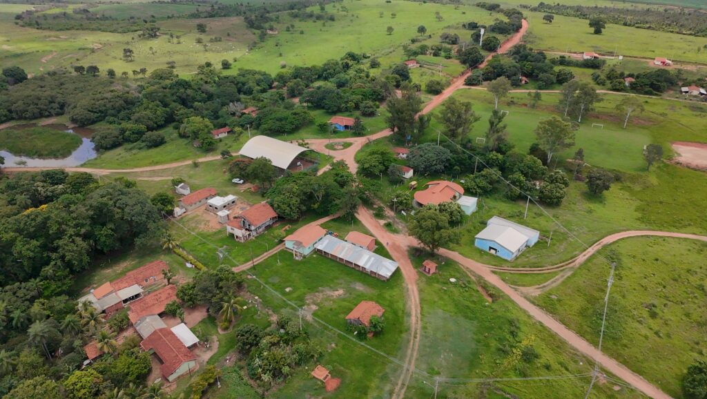 Vista aérea da Terra Indígena Icatu, em Braúna. A imagem mostra uma área ampla com vegetação verde predominante, intercalada por caminhos de terra e pequenas construções distribuídas de forma espaçada. As casas, em sua maioria com telhados claros, estão organizadas ao longo das estradas, cercadas por árvores e áreas abertas. Ao redor da aldeia, há extensões de mata e campos, indicando um território rural e integrado à natureza.