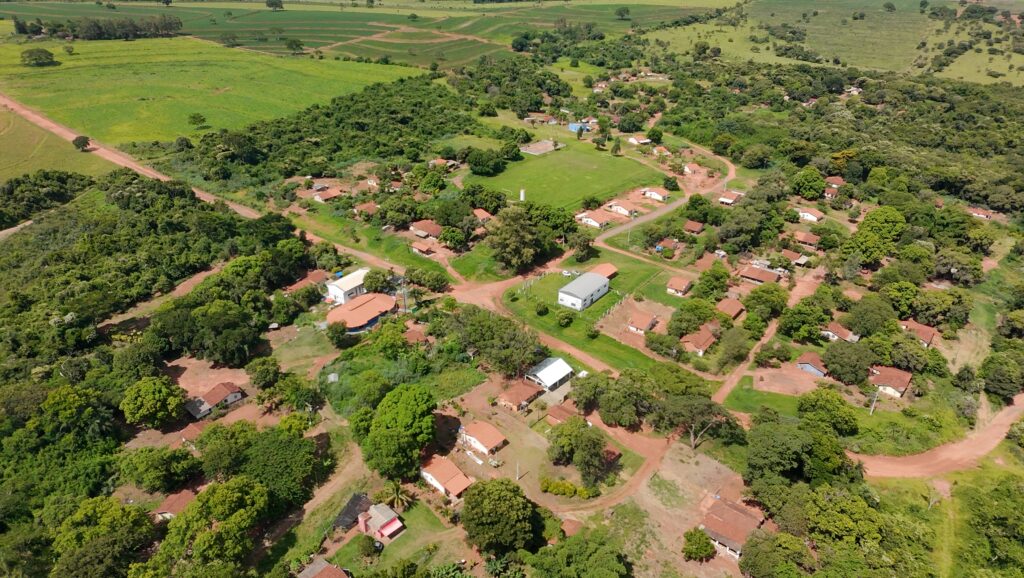 Vista aérea da Terra Indígena Vanuíre, em Arco-Íris. A imagem apresenta uma aldeia com casas distribuídas ao longo de caminhos de terra, cercadas por árvores e áreas verdes. As construções têm telhados claros e estão organizadas de forma espaçada, integradas à paisagem natural. Ao redor, há vegetação densa e áreas abertas, evidenciando um território rural amplo e conectado à natureza.